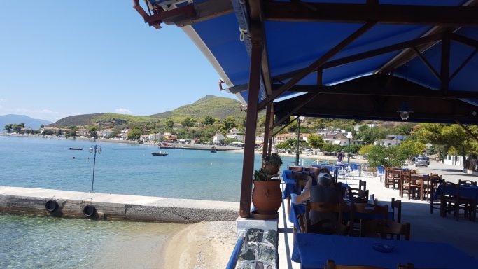 Restaurantterrasse mit blauem Sonnenschirm und Blick auf das Wasser und die umliegenden Hügel.